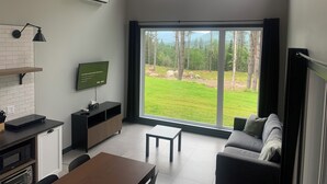 A modern living room with a large sliding glass door leading to a grassy outdoor area, a flat-screen TV mounted on the wall, and a kitchen area with a microwave and a coffee machine. - Valley view north arm#spas#outdoor fireplace#nature#guaranteed fun (Saint-Raymond)