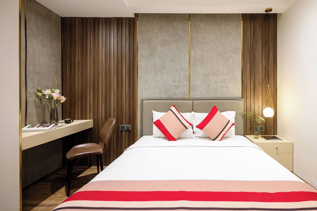 Une chambre d’hôtel moderne comprenant un lit, une table de chevet, une commode, un bureau, une chaise et un vase avec des fleurs.