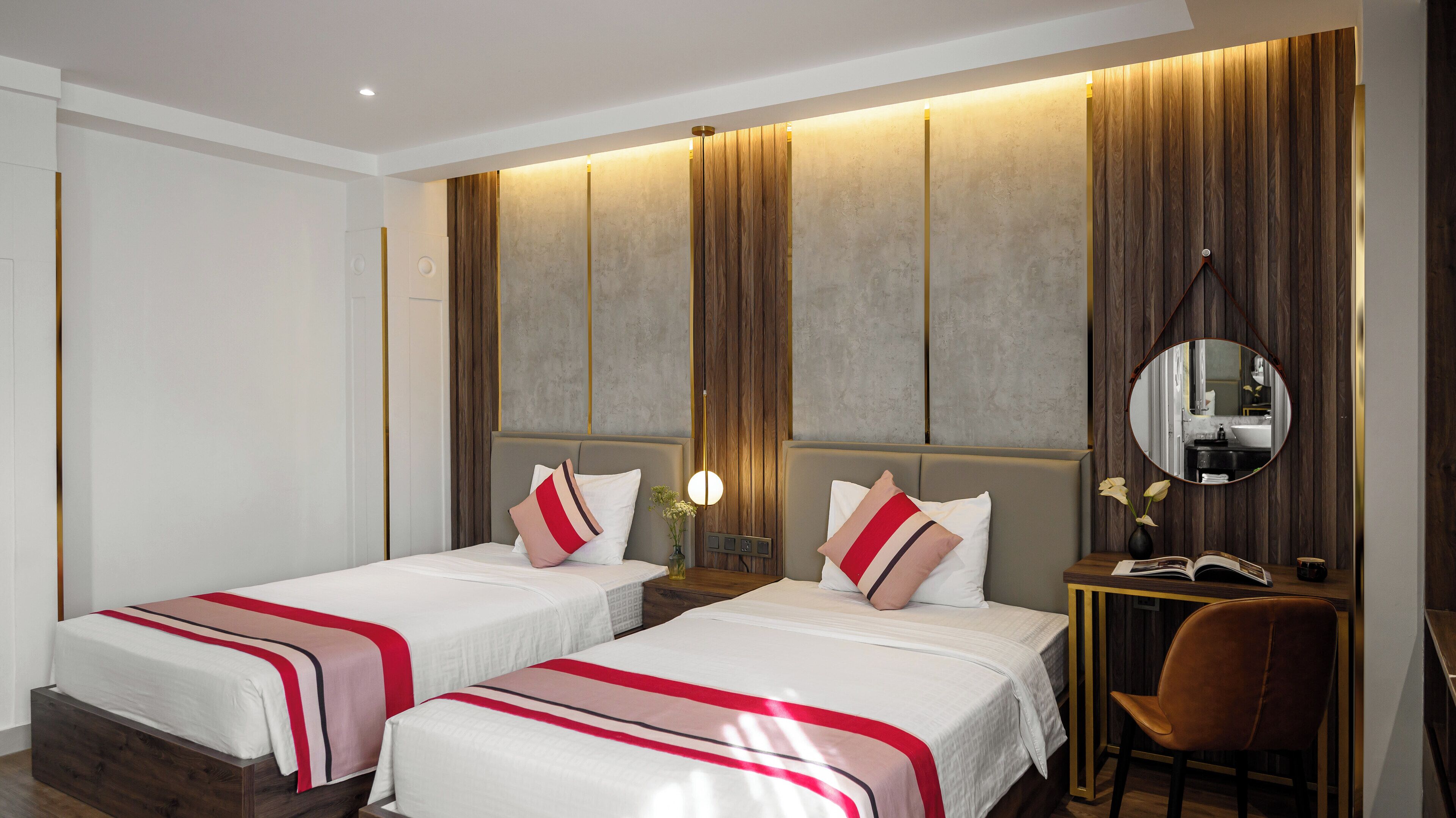 A hotel room with two beds, a desk with a chair, a mirror, and a small table with a vase and book.