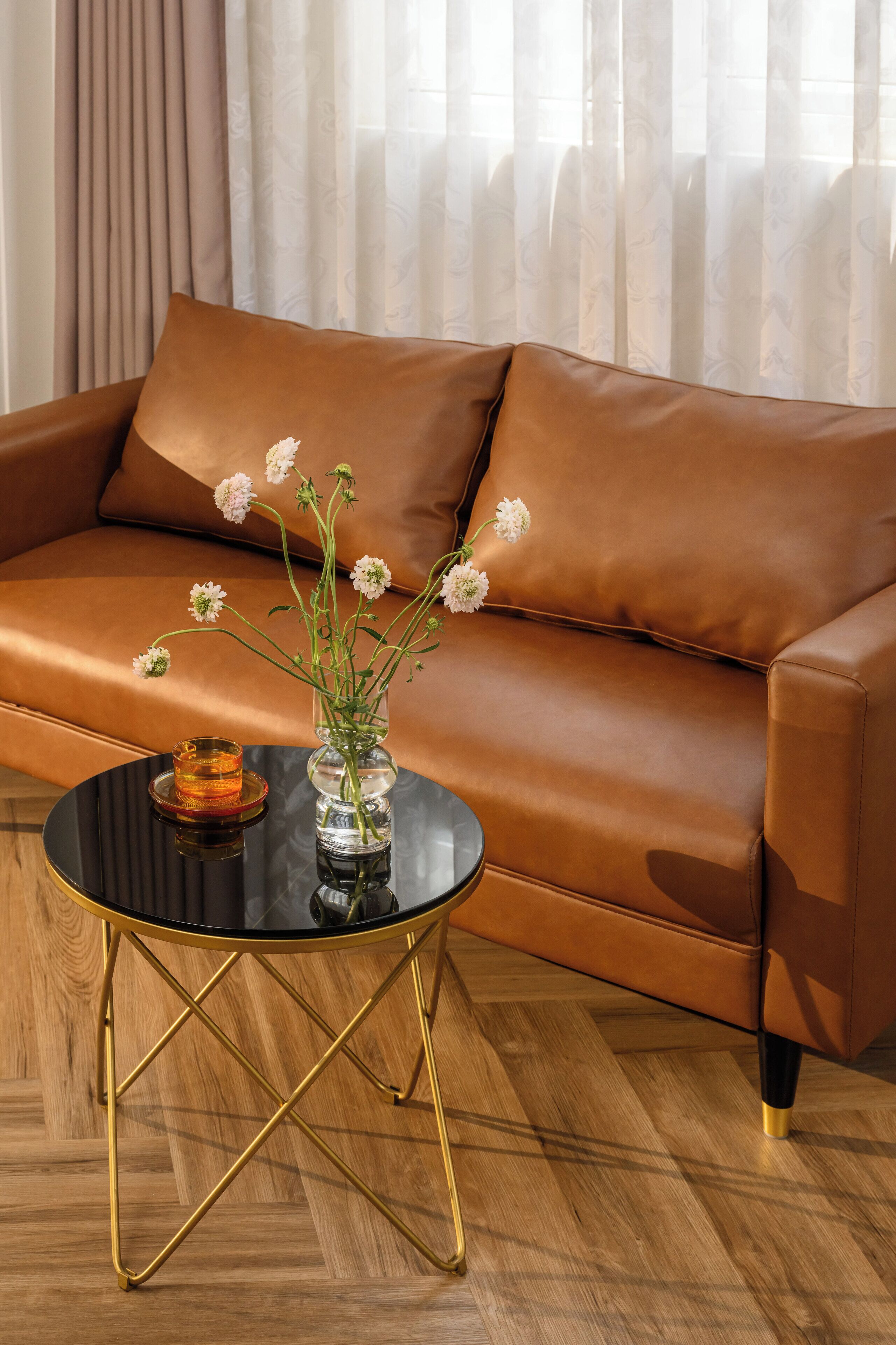 A brown leather sofa with a vase of flowers on a black round table with gold legs.
