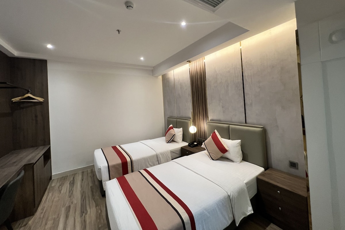 Une chambre d’hôtel avec deux lits, un bureau en bois, un porte-manteau mural et un design moderne au niveau du plafond.