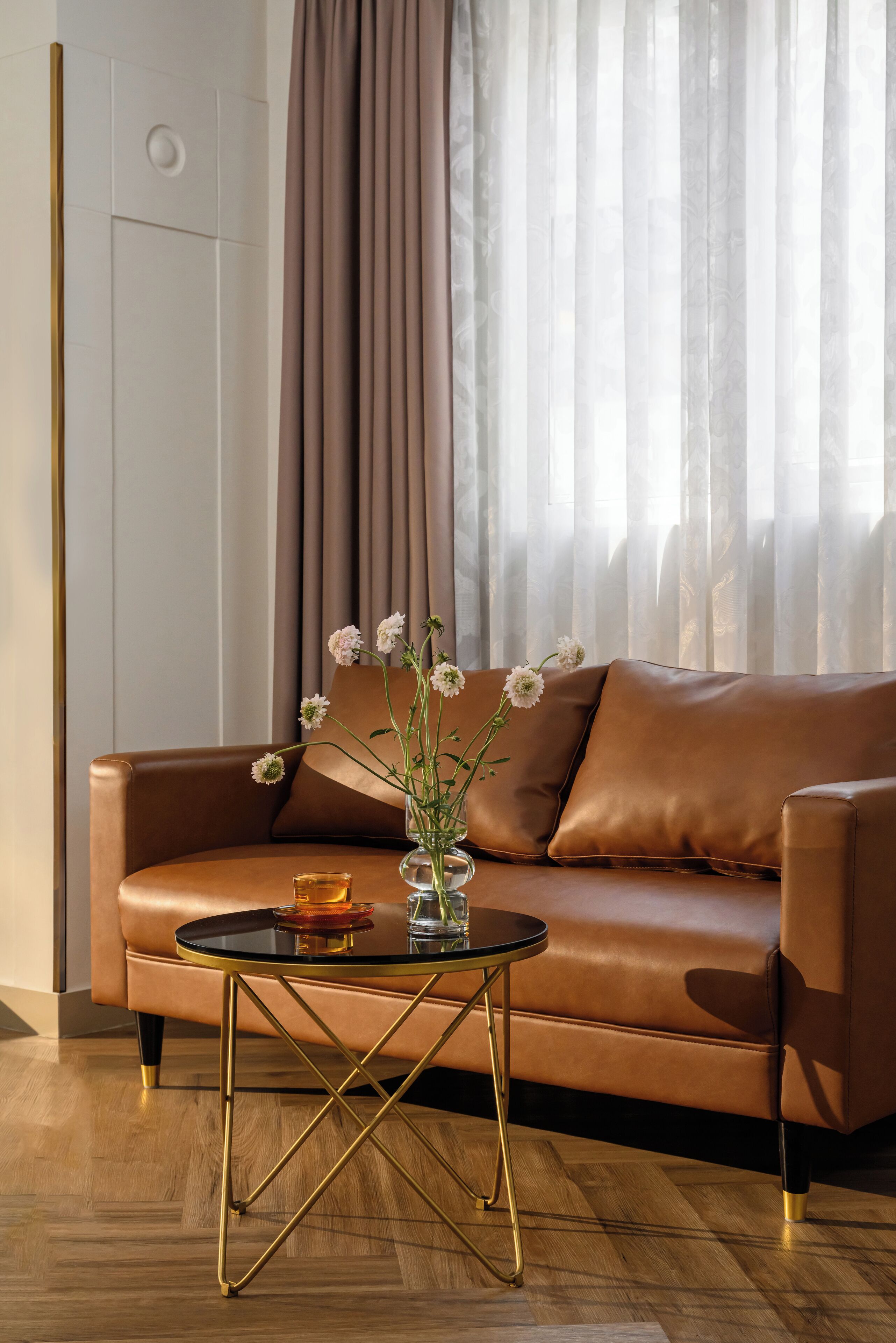 A brown leather sofa with a glass coffee table in front, a vase with flowers, and a window with curtains.