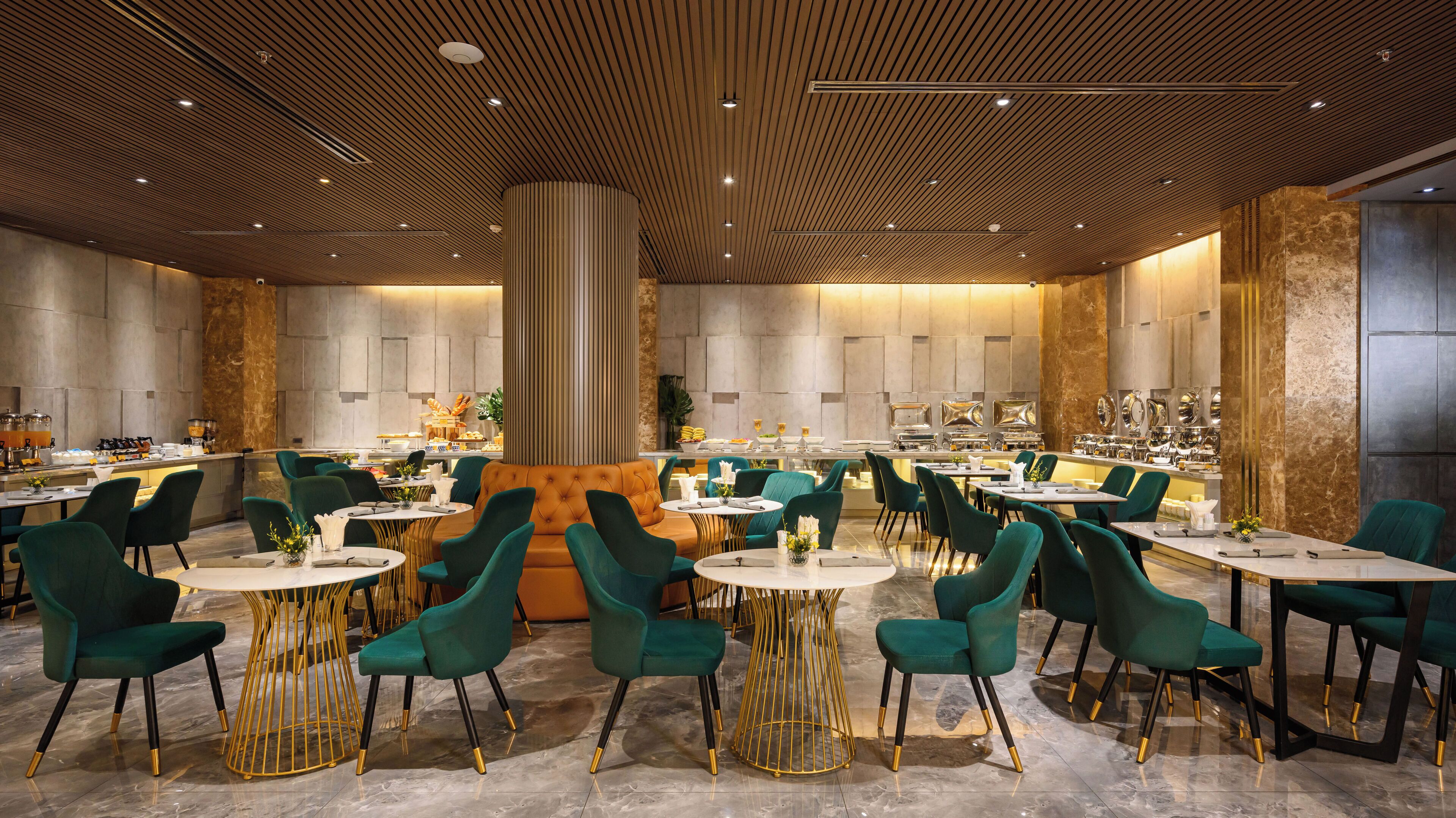 A spacious dining area with round tables and green chairs, a central seating area with a sofa, and a kitchen visible in the background.