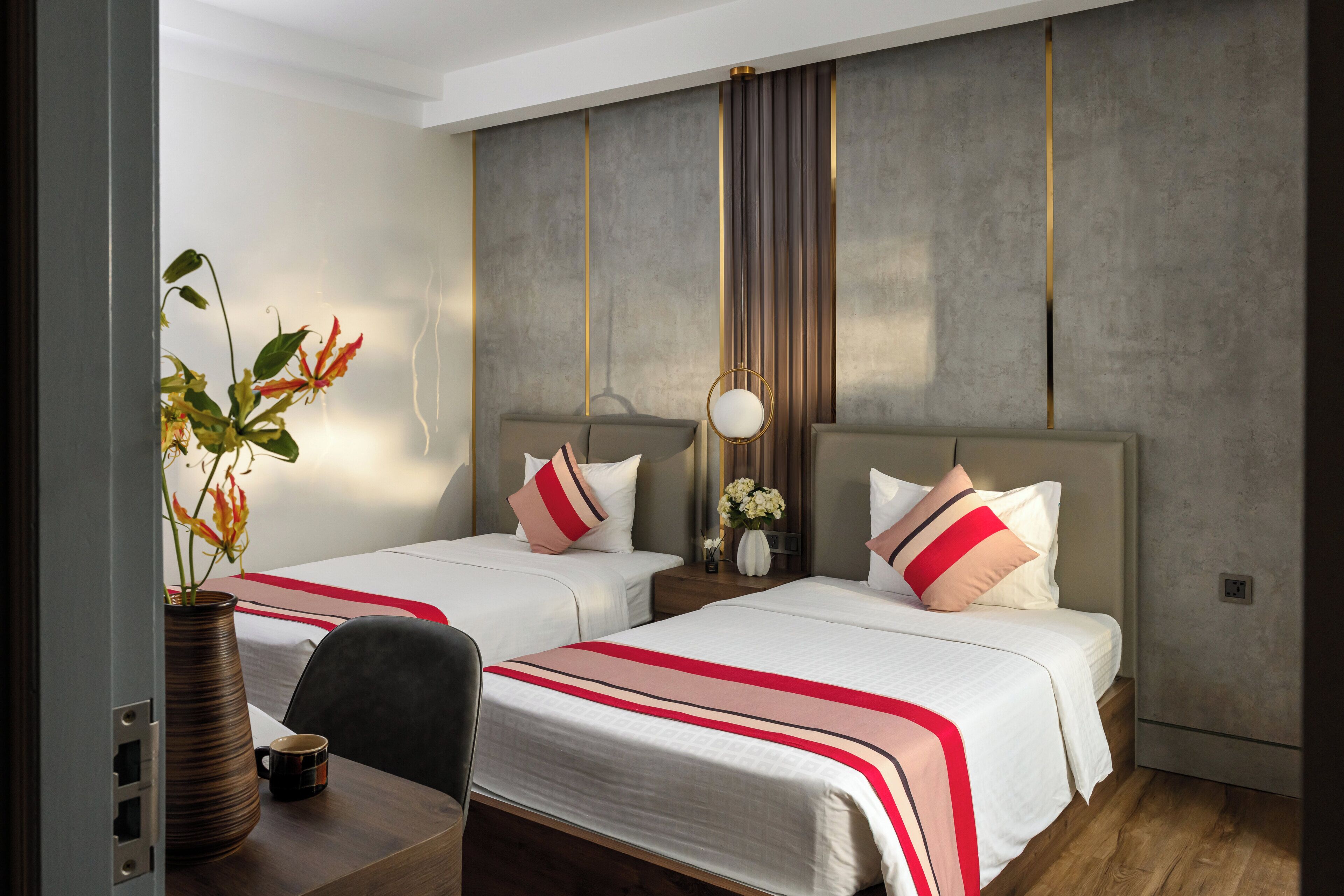 A hotel room with two beds, a desk, a chair, a vase with flowers, and a wall with vertical lines.