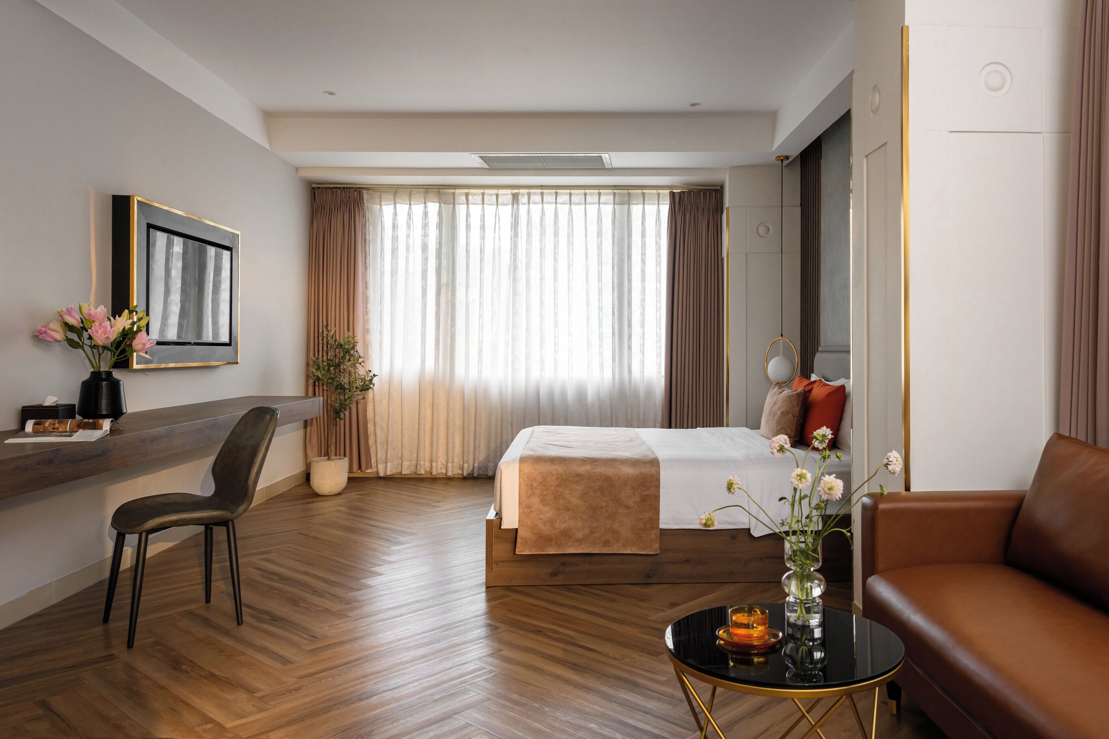 A hotel room with a bed, a sitting area with a couch and a chair, a coffee table, a desk with a chair, a vase with flowers, and a large window with curtains.