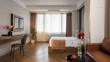 A hotel room with a bed, a sitting area with a couch and a chair, a coffee table, a desk with a chair, a vase with flowers, and a large window with curtains.