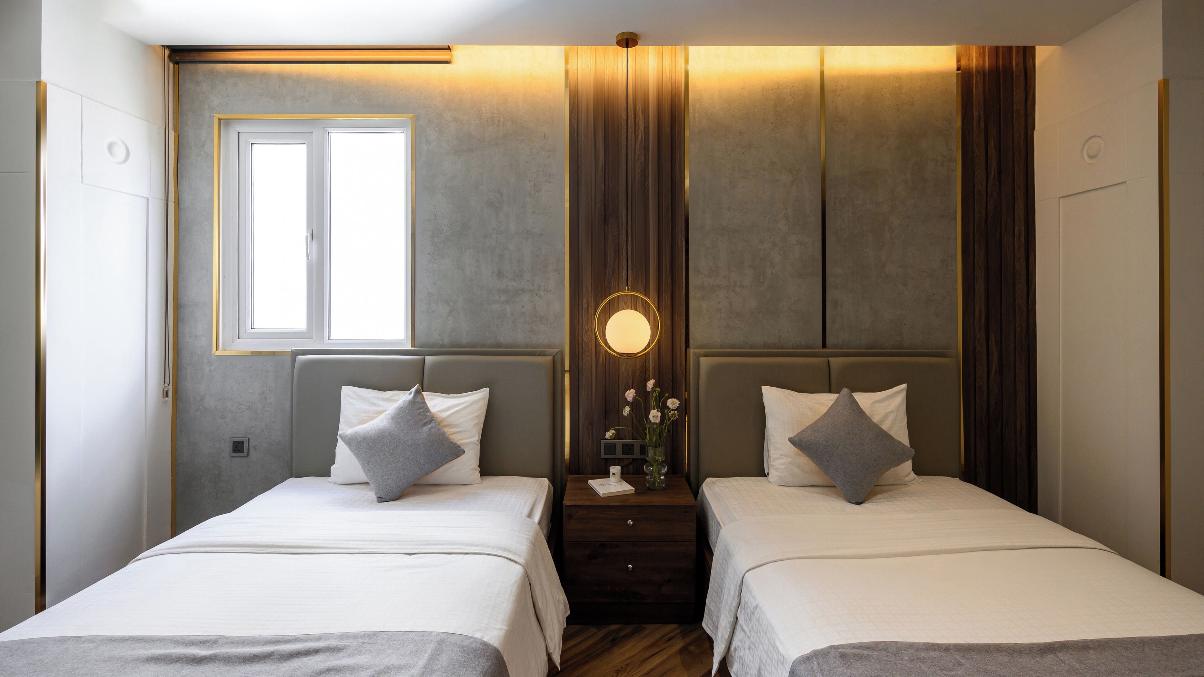 A modern bedroom with two beds, a nightstand with a vase of flowers, and a wall-mounted light fixture.