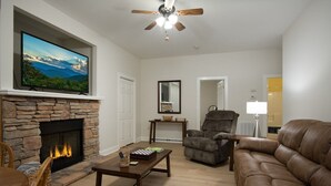 A living room with a stone fireplace, a flat-screen TV, a leather sofa, and a ceiling fan. - Hummingbird Hideaway in Connestee Falls (Brevard)