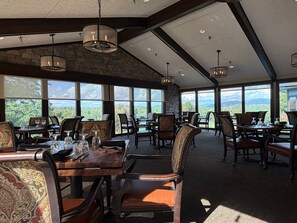 A dining area with tables and chairs, large windows, and a stone wall.
