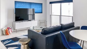 Un salon moderne avec un canapé en cuir noir, une table basse en verre, une table à manger blanche avec des chaises bleues, un téléviseur posé sur un meuble blanc et une grande fenêtre avec des stores bleus.