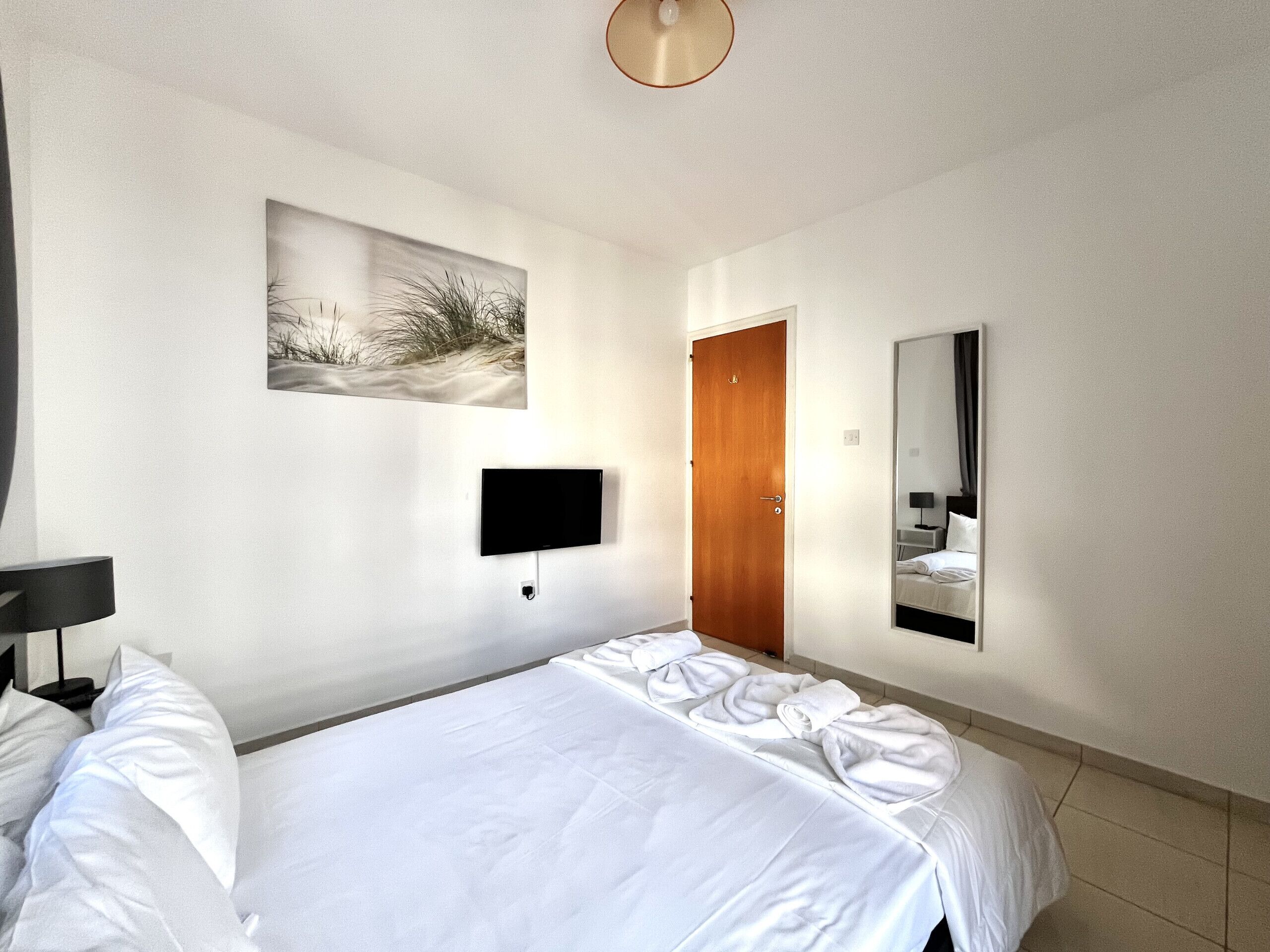 A hotel room with a bed, a flat-screen TV mounted on the wall, a wooden door, and a large mirror.