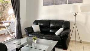 Ein modernes Wohnzimmer mit einem schwarzen Ledersofa, einem Glastisch und einer Stehlampe. An der Wand hängen drei gerahmte Bilder und auf dem Tisch steht eine kleine Pflanze.