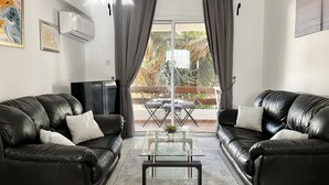 Ein Wohnzimmer mit zwei schwarzen Ledersofas, einem Glastisch und einer großen Glasschiebetür, die zu einem Außenbereich mit Tisch und Stühlen führt.