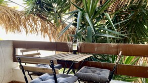 Ein Sitzbereich im Freien mit einem Holztisch und zwei Klappstühlen, umgeben von Pflanzen und einem Reetdach.