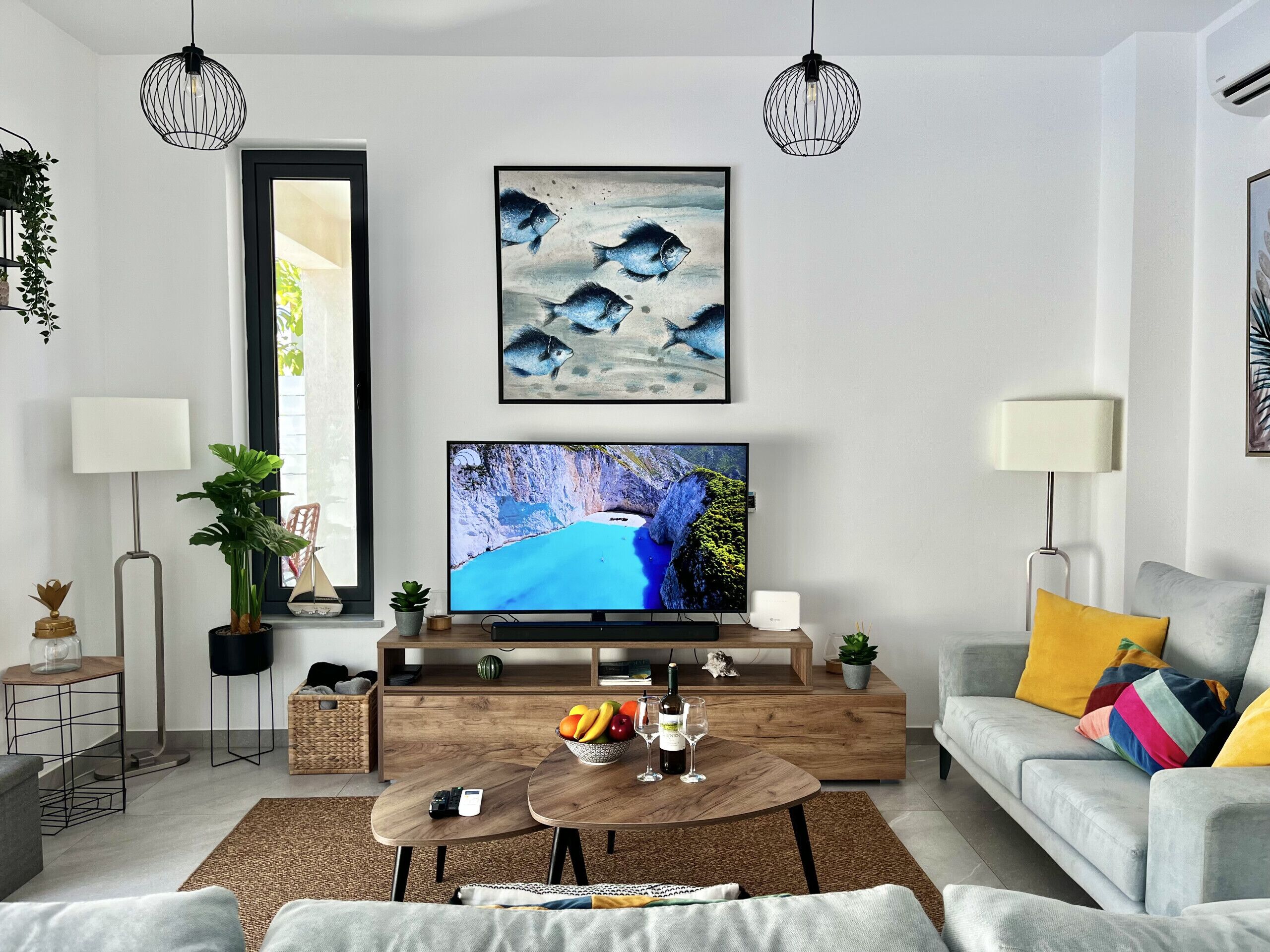 En moderne stue med et fladskærms-tv, et træsofabord, en grå sofa og dekorative planter.