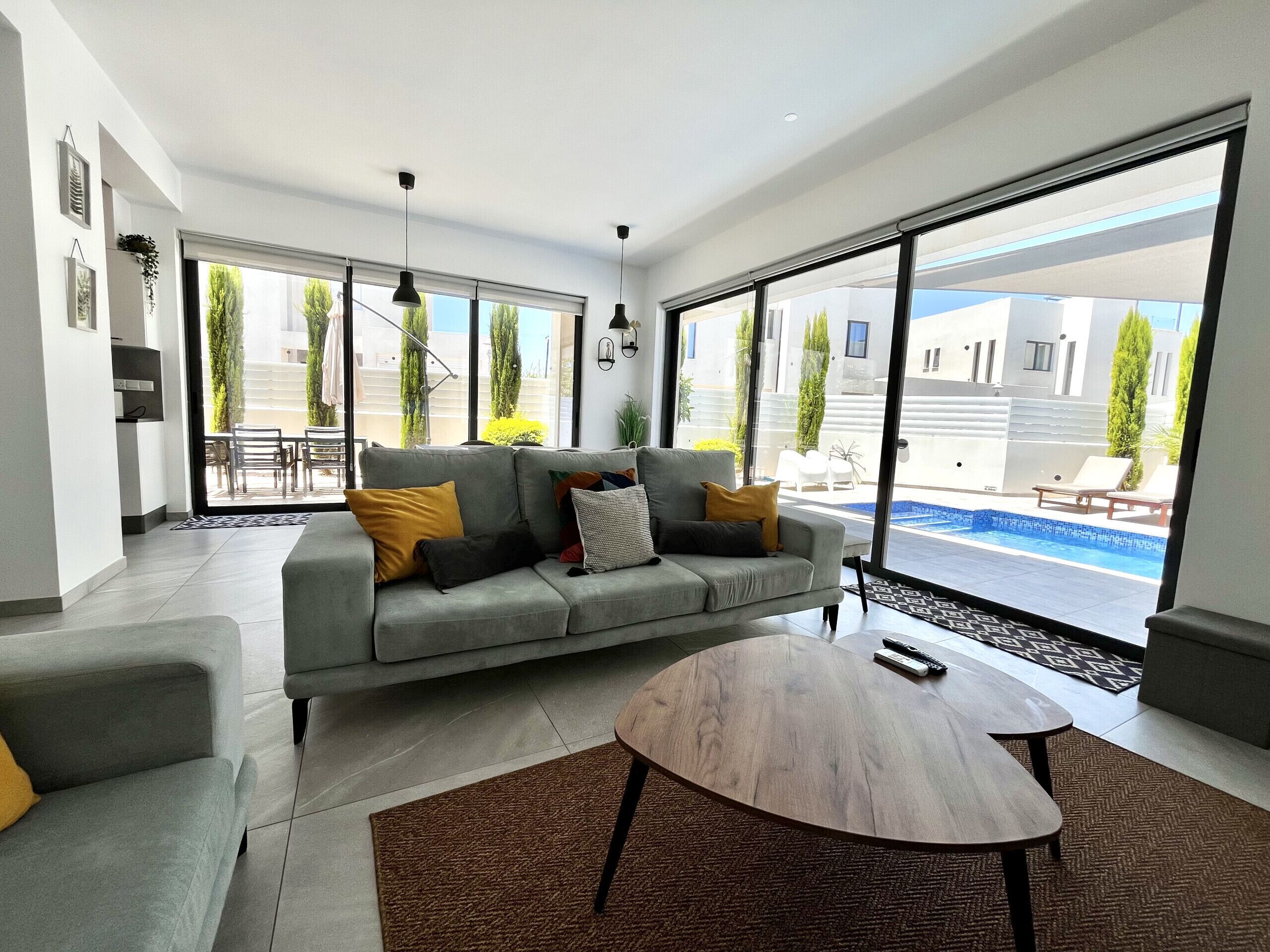 A modern living room with a grey sofa, a round wooden coffee table, and large sliding glass doors leading to an outdoor pool area.