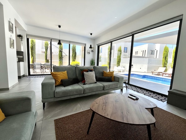 A modern living room with a grey sofa, a round wooden coffee table, and large sliding glass doors leading to an outdoor pool area.