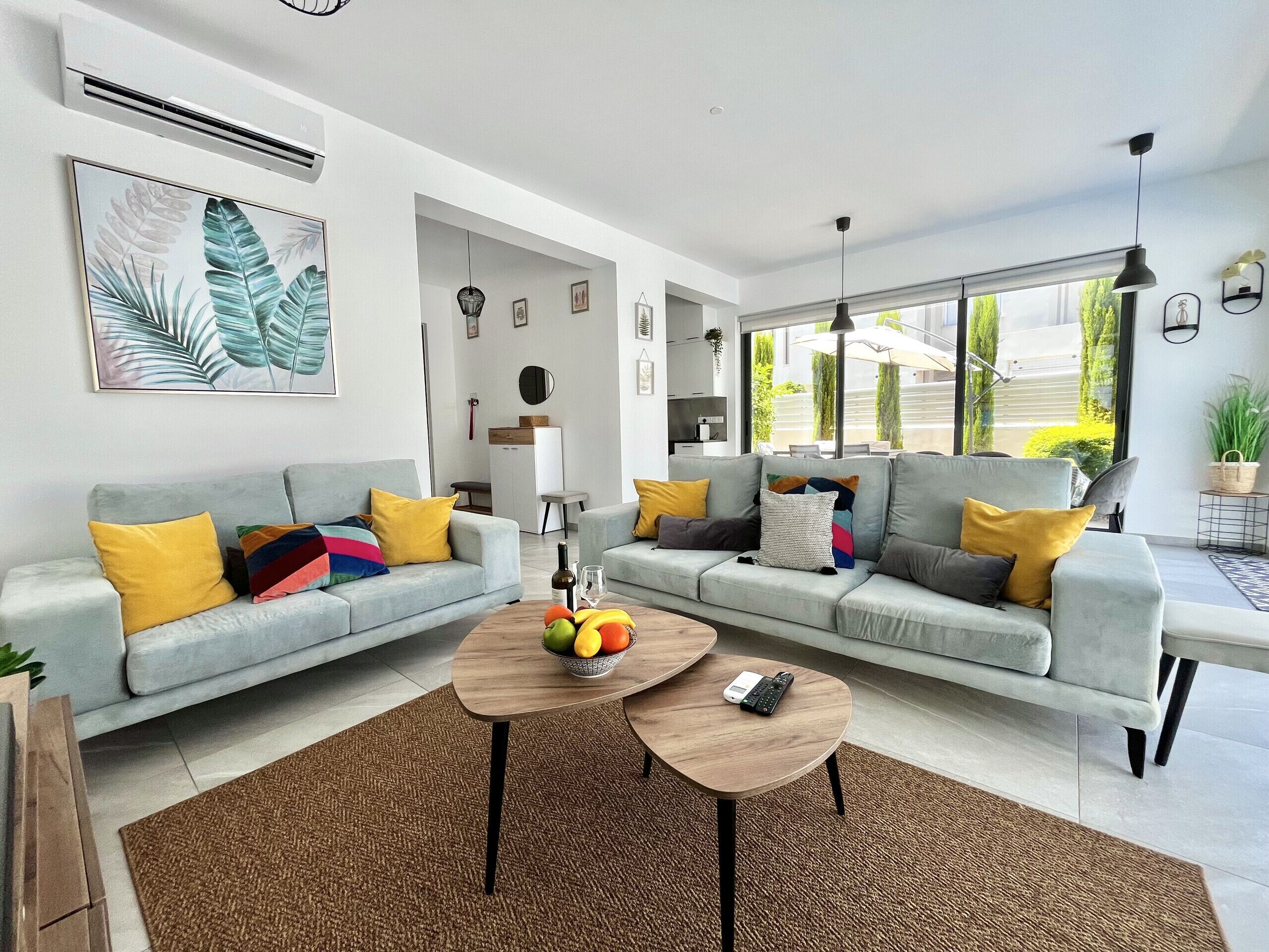 A modern living room with two grey sofas, colorful cushions, a coffee table with fruits, and a large window offering a view of greenery.