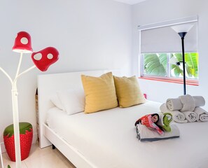 Een net opgemaakt bed met witte en gele kussens, een rode decoratie op een witte standaard, en een raam met uitzicht op groene begroeiing.