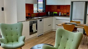 A modern kitchen with white cabinets, a stainless steel stove, and a white refrigerator. There are two green armchairs and a small table in the dining area.
