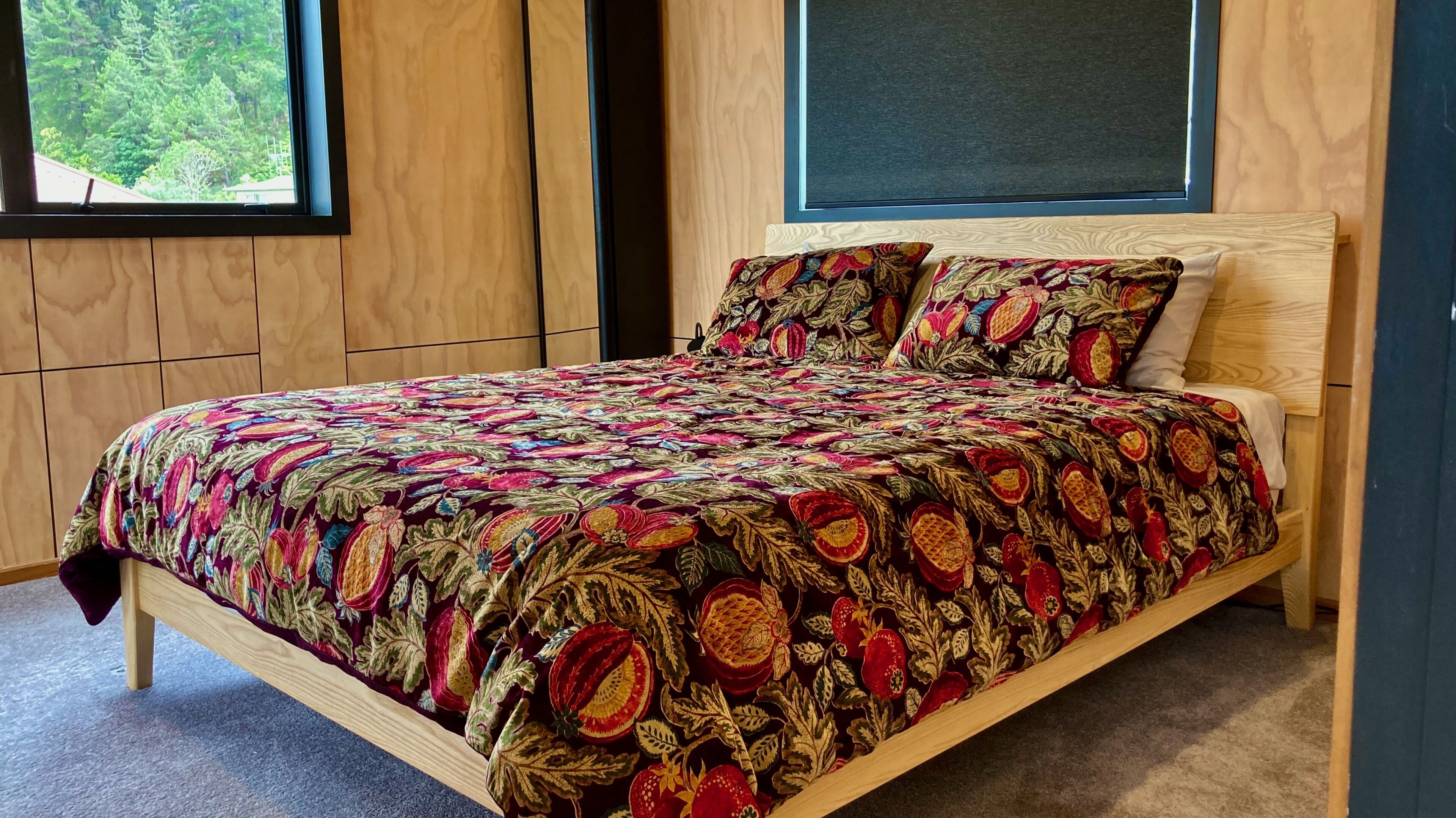 Un lit en bois, recouvert d’une couvre-lit à motifs floraux et agrémenté d’oreillers, une fenêtre donnant sur des arbres et un mur sombre derrière le lit.