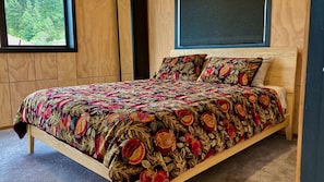 A wooden bed with a floral-patterned bedspread and pillows, a window with a view of trees, and a dark wall behind the bed.