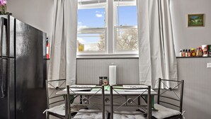 A kitchen with a black refrigerator, a dining table with four chairs, a window with curtains, and a shelf with condiments.