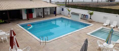 Zona de piscina con dos piscinas, sillas de descanso y un área de estar cubierta.