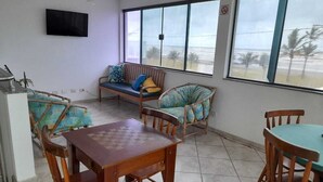 A room with a wooden table and chairs, a sofa with cushions, and large windows overlooking a beach. - CASABLANCA Praia Hotel (Peruíbe)