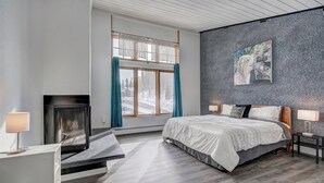 A modern bedroom with a fireplace, a large bed, a painting on the wall, and a view of a snowy landscape through the window. - 3bd_cj_12 (Breckenridge)
