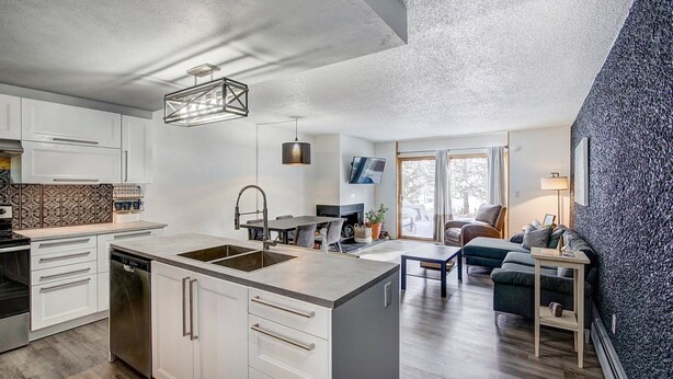 A modern kitchen with white cabinetry, a central island with a sink, and a dining area with a table and chairs. - 3bd_cj_12 (Breckenridge)