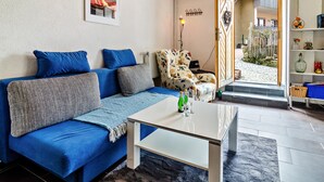 A living room with a blue sofa, a white coffee table, a patterned armchair, and a doorway leading to a courtyard.