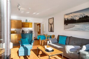 Ein modernes Wohnzimmer mit Sofa, Sessel und Couchtisch, eine Küche mit Holzschränken und eine Essecke mit Tisch und Stühlen.