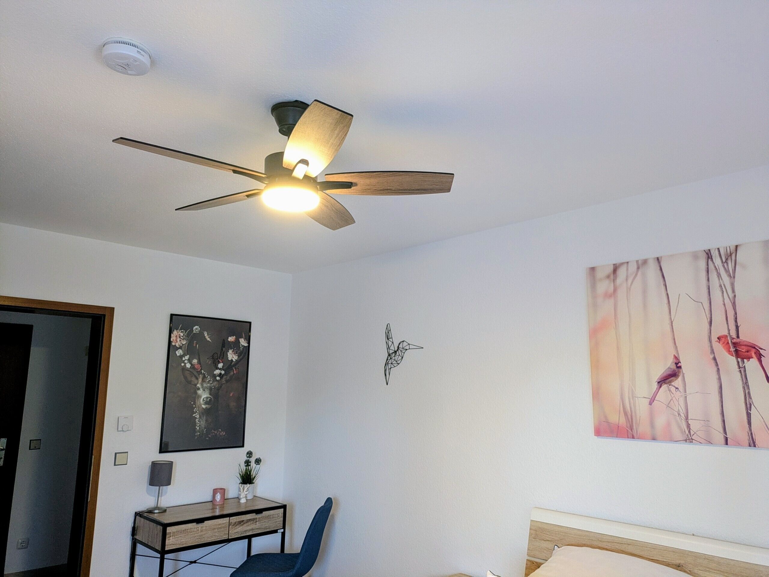 Ein Schlafzimmer mit Deckenventilator, einem Bett, einem Schreibtisch mit Stuhl und einem Wandbild.