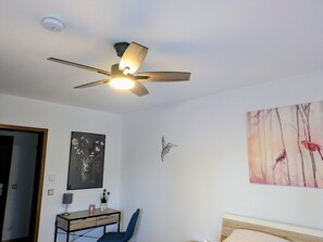 Une chambre dotée d’un ventilateur de plafond, d’un lit, d’un bureau avec une chaise et d’une œuvre d’art accrochée au mur.