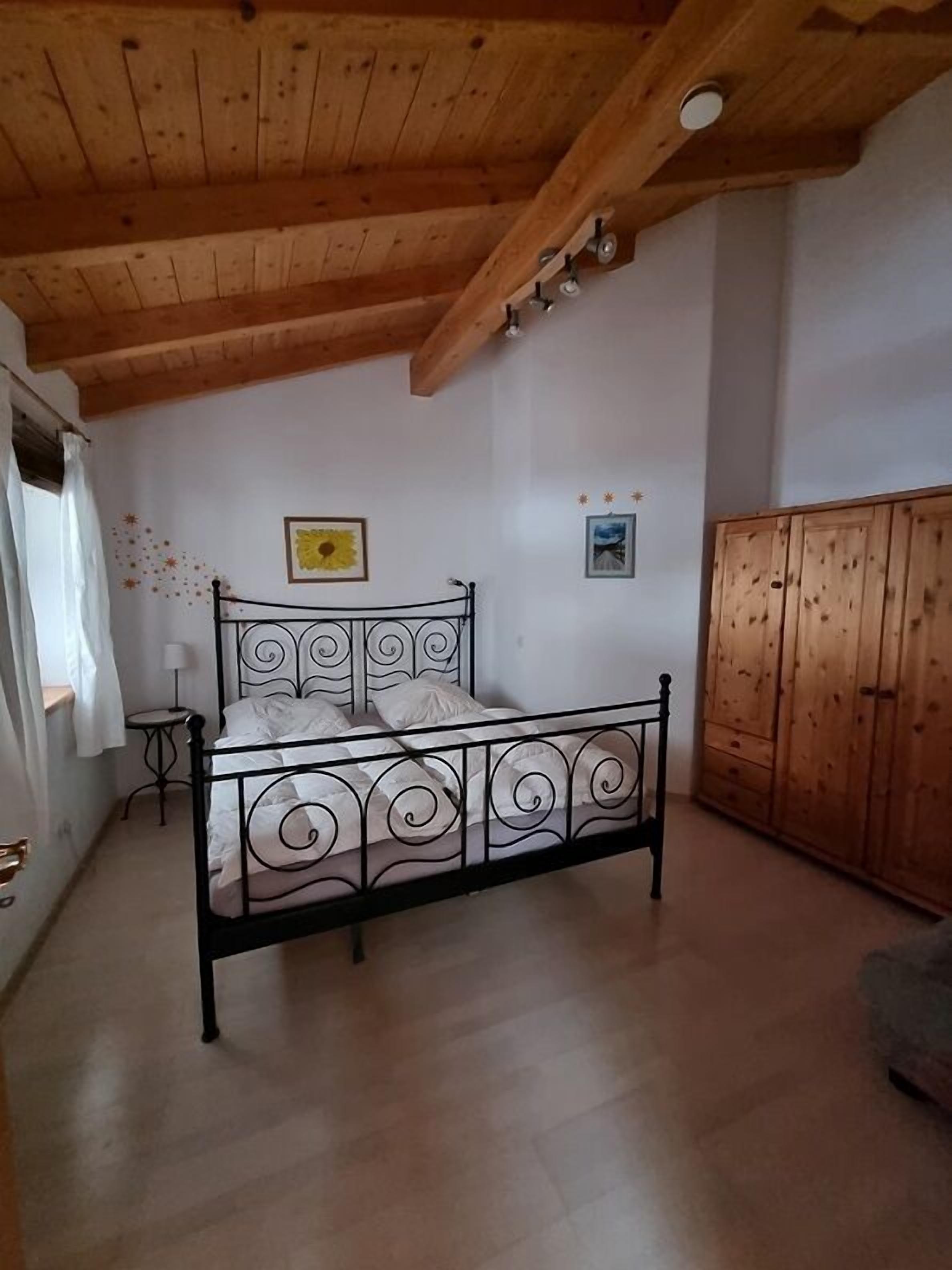 A bedroom with a metal bed frame, wooden ceiling beams, a wardrobe, and a small bedside table.