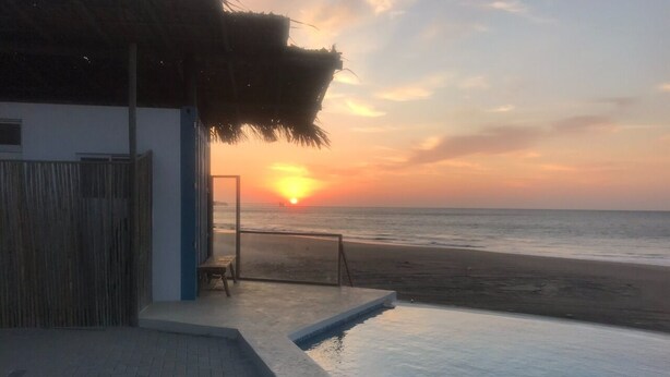 A swimming pool with a view of the sunset over the ocean, a wooden fence, and a thatched roof structure. - COCOBOX (Los Organos)