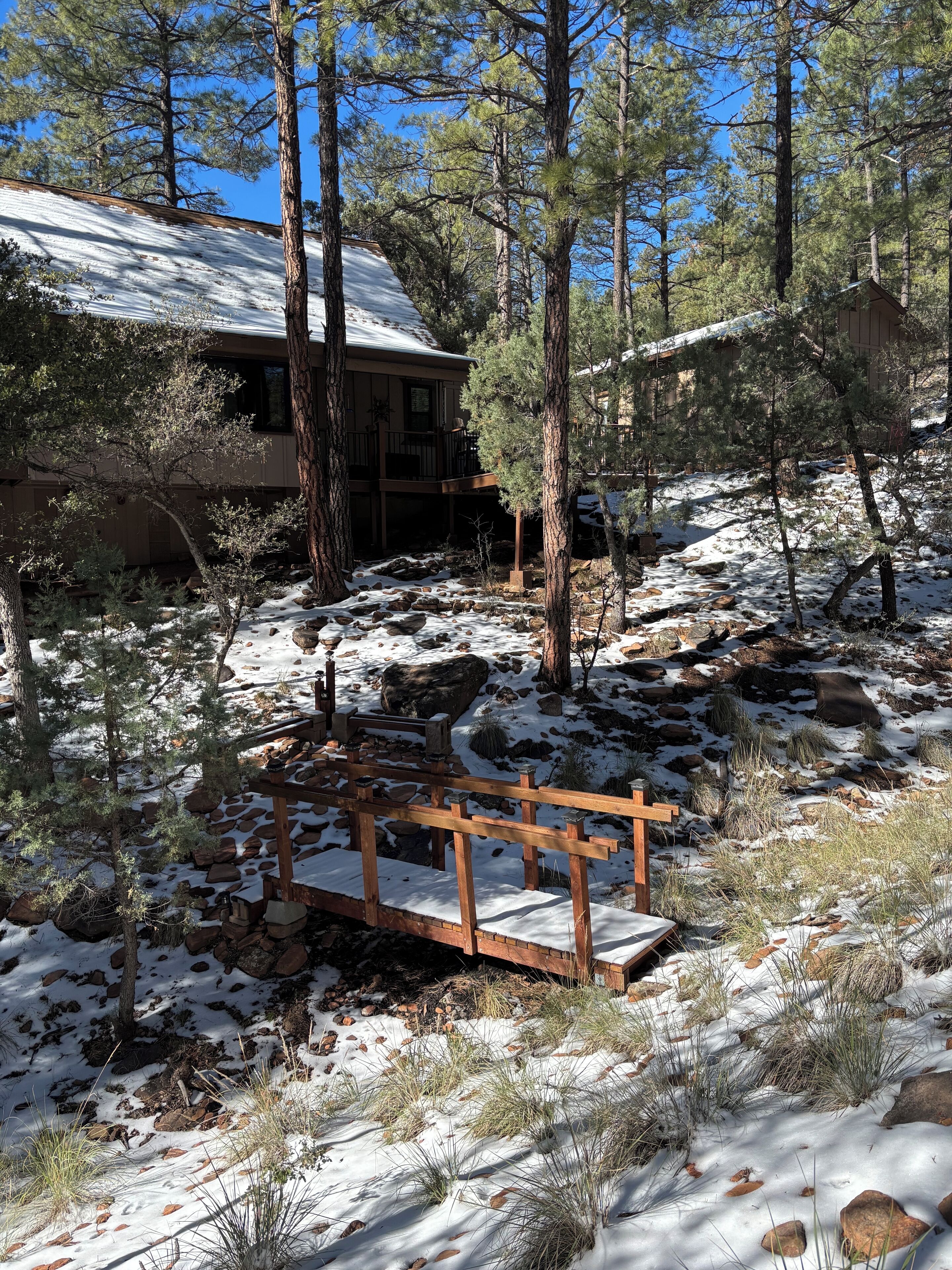 Une cabane dotée d’une terrasse en bois et d’un pont de pierre, située dans une forêt enneigée.