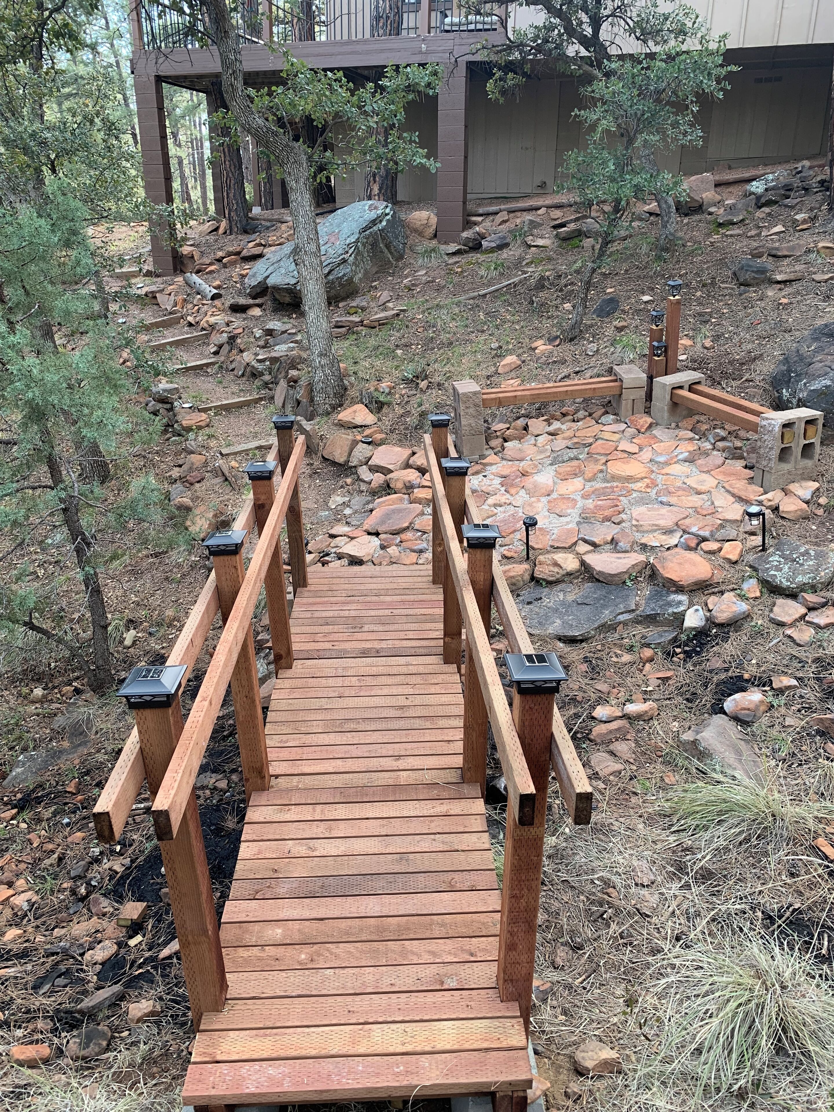 A wooden bridge with railings leading to a stone patio area surrounded by trees and rocks.