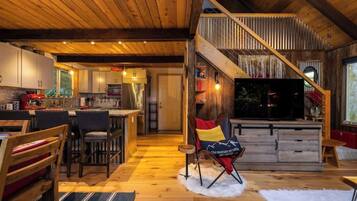 A spacious living area with a wooden ceiling, a dining area with chairs, a kitchen with cabinets, and a TV in the living room.