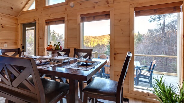Eine Holzhütte mit Essbereich, einem Tisch für vier Personen, großen Fenstern und Blick auf den umliegenden Wald.