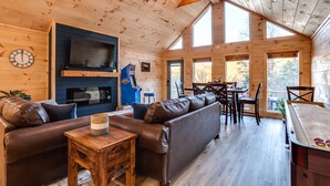 Ein gemütliches Blockhaus-Interieur mit Holzbalkendecke, Kamin, Ledersofas, Essbereich und großen Fenstern.