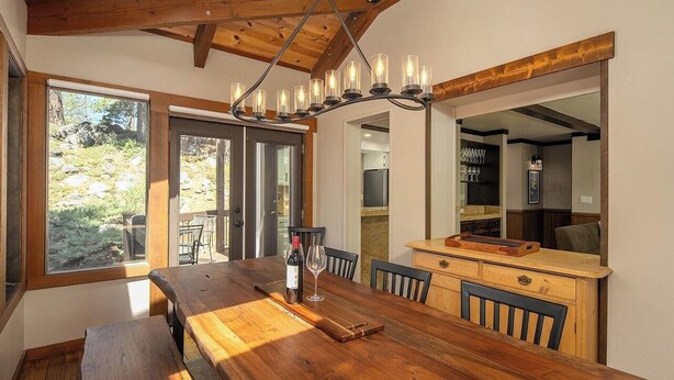 Een houten eettafel met stoelen, een fles wijn en een glas. Daarboven een kroonluchter en aan de muur een grote spiegel.