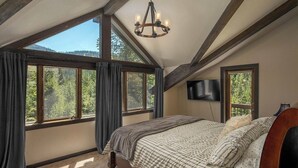 Een slaapkamer met een groot bed, een kroonluchter en een televisie. Er zijn grote ramen met gordijnen, en uitzicht op bomen en heuvels.