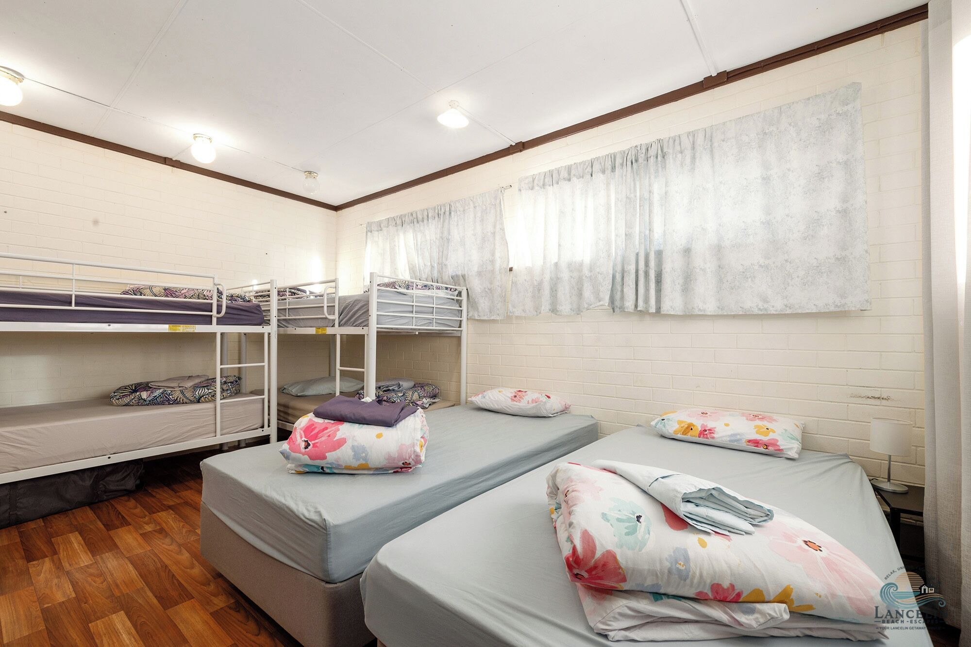 A dormitory room with bunk beds, a window with curtains, and a wooden floor.
