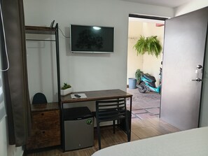 A compact room with a bed, a desk with a chair, a small refrigerator, a TV mounted on the wall, and a door leading outside.