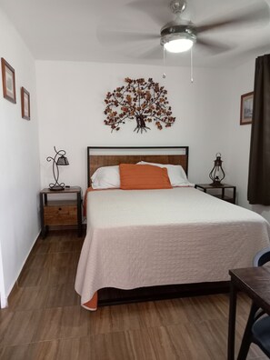 A neatly arranged bedroom with a bed, a nightstand, a lamp, a ceiling fan, and a wall-mounted tree decoration.