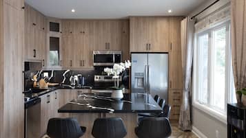 Cozinha moderna com armários de madeira, ilha central com banquetas, eletrodomésticos de aço inoxidável e uma grande janela com cortinas.