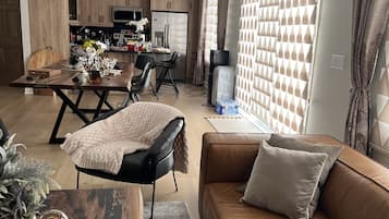 Un salon moderne avec un canapé en cuir marron, une table à manger en bois et un coin cuisine à l’arrière-plan.