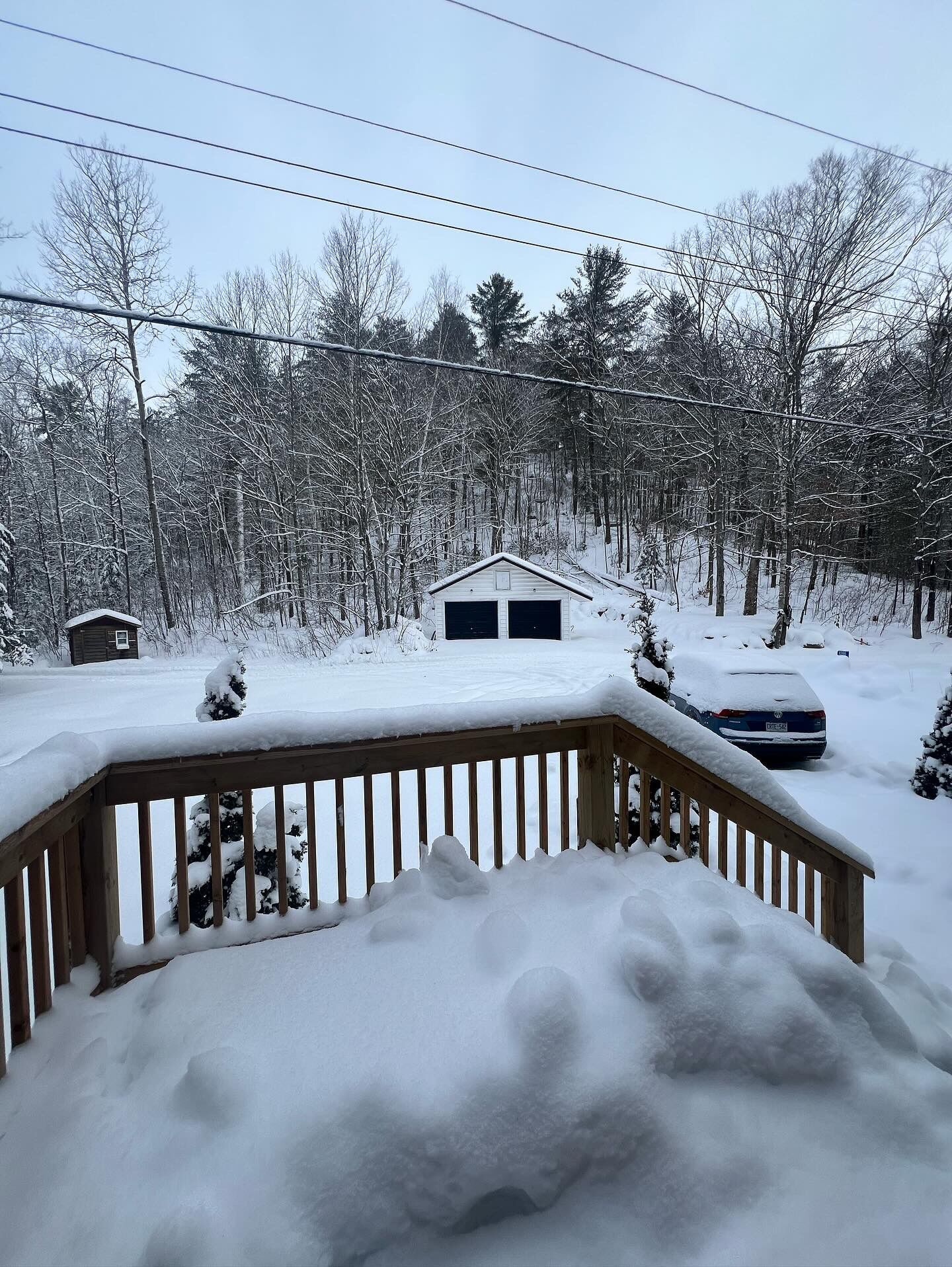 A snow-covered deck with a wooden railing, a small shed, and a car parked in the background.